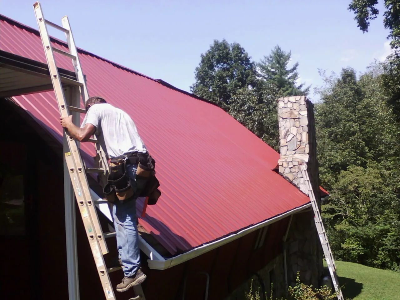 Roofer on a ladder beside a red metal roof and stone chimney — Soffit & Fascia Repair by Roofing Brothers in Harbor View